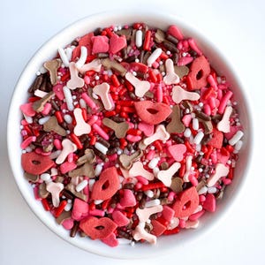 May include: A white bowl filled with colorful sprinkles, including red, pink, brown, and white. The sprinkles are shaped like hearts, lips, and other abstract shapes.