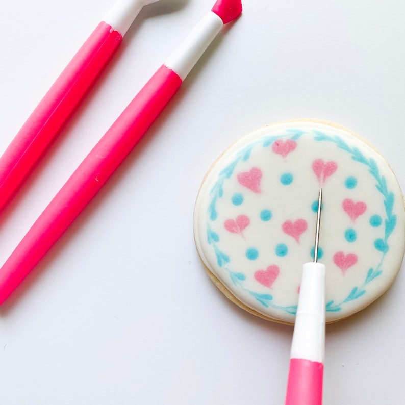 May include: A pink cookie decorating tool with a white tip is being used to create a design on a round sugar cookie. The cookie is decorated with a blue and pink floral pattern.