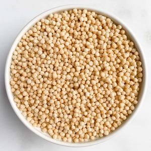 May include: A white bowl filled with small, round, beige-colored couscous.