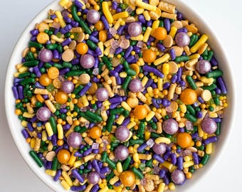 May include: A bowl of colorful sprinkles in green, yellow, purple, and gold. The sprinkles are a mix of small balls, stars, and long, thin pieces.