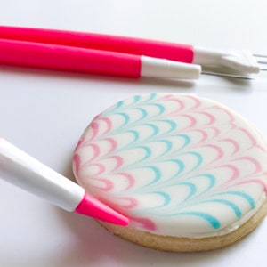 May include: A white cookie with pink and blue icing in a wave pattern is being decorated with a pink icing pen.