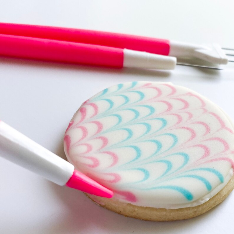 May include: A white cookie with pink and blue icing in a wave pattern is being decorated with a pink icing pen.