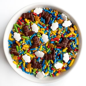 May include: A bowl of colorful sprinkles with a cowboy theme. The sprinkles include brown boots, white clouds, yellow stars, and red, blue, green, and yellow sprinkles.