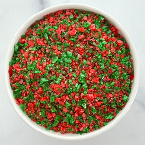 May include: A white bowl filled with red and green crushed candy. The candy pieces are irregularly shaped and have a glossy appearance. The bowl is set against a white background.