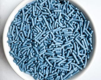 May include: A white ceramic bowl filled with blue sprinkles. The sprinkles are small, cylindrical, and evenly distributed throughout the bowl. The bowl is round with a slightly raised edge, and the background is a plain white surface.