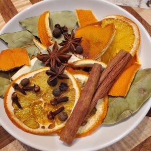 May include: A white bowl filled with dried orange slices, star anise, cinnamon sticks, bay leaves, cloves, and peppercorns. The ingredients are arranged on a wooden surface, creating a warm, inviting aesthetic. Ideal for potpourri or festive decor.