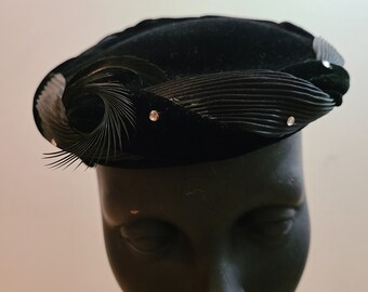 May include: A black velvet hat with a decorative feather and rhinestone embellishment. The hat has a rounded shape with a curled feather design on the side. The hat is displayed on a mannequin head.