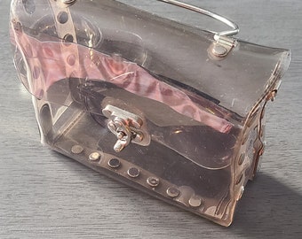 May include: A clear, rectangular handbag with a silver handle and clasp. The transparent material reveals a pinkish interior. Silver rivets secure the sides. The bag sits on a gray surface.