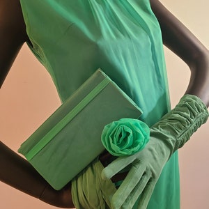 May include: A vintage green sleeveless dress with a matching clutch, gloves, and a floral accent. The dress features a high neckline and a delicate floral embellishment. The gloves are long and ruched, and the clutch is rectangular.