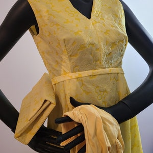 May include: A sleeveless, yellow floral-patterned dress with a V-neck, a matching clutch, and gloves. The dress has a fitted bodice and a slightly flared skirt. The gloves are a light yellow color. The outfit is displayed on a black mannequin.