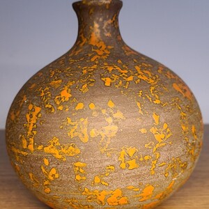 May include: A brown ceramic vase with a rounded body and a narrow neck. The vase is adorned with a textured, speckled pattern in shades of orange and brown. The vase is displayed on a wooden surface.