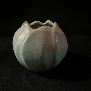 May include: A small, round, green and gray ceramic vase with a scalloped rim. The vase has a textured surface and a subtle ombre effect.