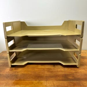 May include: A stackable, beige-colored desk organizer with three trays. Each tray has a rectangular shape with a slightly raised front and a cutout on the sides for easy access. The organizer is made of a durable plastic material.