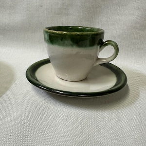 May include: A white ceramic teacup with a green rim and a matching saucer. The cup has a rounded shape and a handle.