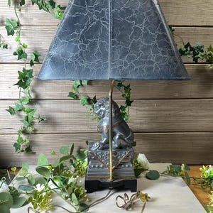 May include: A decorative table lamp with a gray, crackled shade and a detailed lion statue base. The lamp has a gold-colored stem and a black base. The cord is visible.