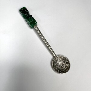 May include: A decorative spoon with a green jade handle and a silver-colored bowl. The bowl features an engraved circular design. The handle is carved with a stepped design. The spoon is set against a white background.