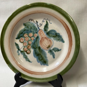 May include: A white ceramic plate with a green, brown, and blue hand-painted design of a pear and grapes. The plate has a green and brown rim.