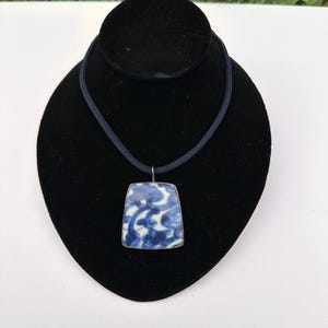 May include: A blue and white porcelain pendant with a floral design hangs from a black cord necklace.