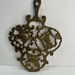 May include: A brass trivet with an intricate design featuring three stylized animals. The trivet has a loop at the top for hanging.