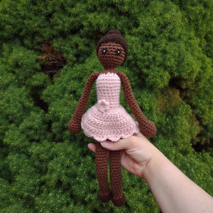 May include: A crocheted doll with brown skin, black eyes, and a pink dress. The doll is being held in a hand.