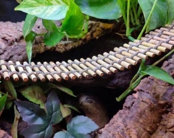 May include: A flexible bridge constructed from numerous small, light-colored wooden dowels connected by thick, dark-toned cord. The bridge is positioned in a curved shape, resting between pieces of textured brown bark and green foliage.