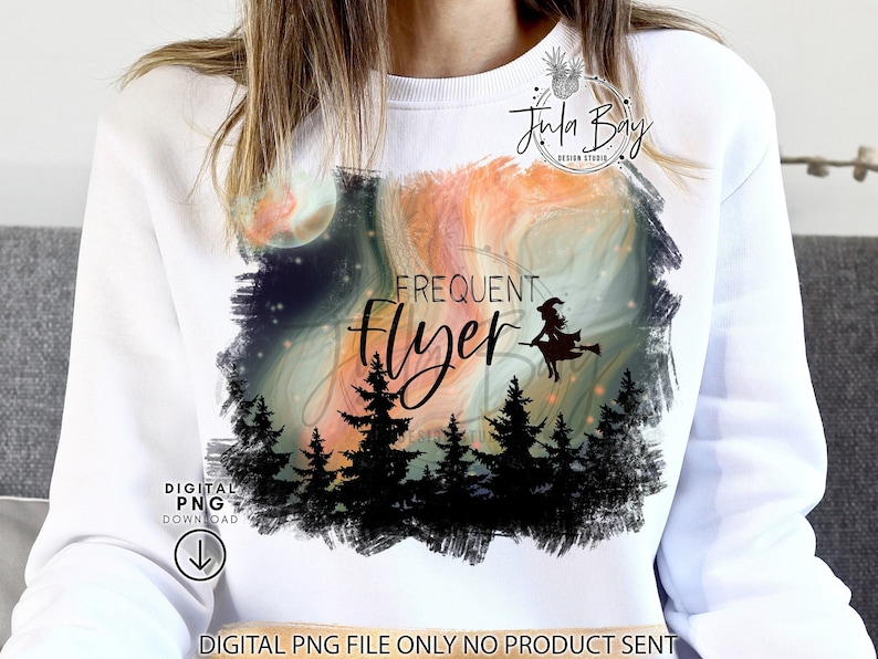 May include: A white crewneck sweatshirt featuring a graphic print of a silhouette of a witch on a broomstick flying over a forest of pine trees against a swirling orange and dark blue sky with the text Frequent Flyer in black lettering.