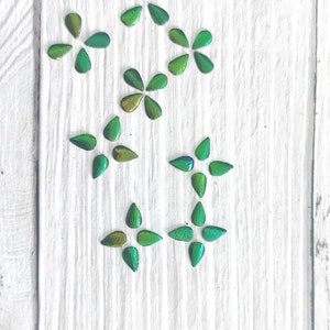 May include: A collection of green and gold iridescent flower-shaped embellishments arranged on a white wooden surface.