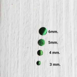 Puede incluir: Cuatro círculos iridiscentes verdes de diferentes tamaños sobre un fondo de madera blanca. Los círculos están etiquetados con su diámetro en milímetros: 6 mm, 5 mm, 4 mm y 3 mm.