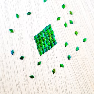 May include: A green iridescent square made of small squares, surrounded by smaller green iridescent diamonds on a white wood surface.