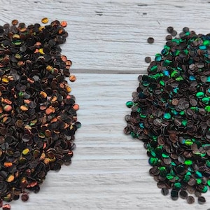 May include: Two piles of iridescent glitter confetti. One pile is black with a copper sheen, the other is black with a green sheen.