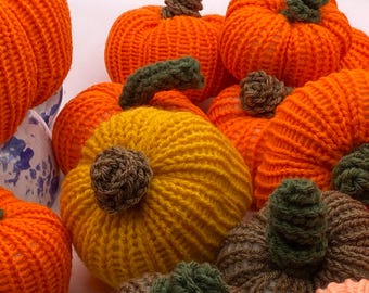 May include: A collection of handmade knitted pumpkins in various shades of orange and yellow. Each pumpkin features a brown or green stem, creating a cozy, autumnal theme. The pumpkins are arranged together, suggesting a fall harvest display.