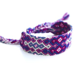 以下が含まれることがあります： 青、ピンク、白の色合いの手作りのフレンドシップブレスレット。織り込まれたブレスレットは幾何学模様で、留め具には編み込みの紐が付いています。毎日の装いにぴったりのカラフルなアクセサリーです。