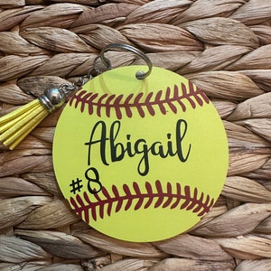 May include: Yellow softball keychain with red stitching and the name Abigail and number 8 printed on it. The keychain has a yellow tassel.