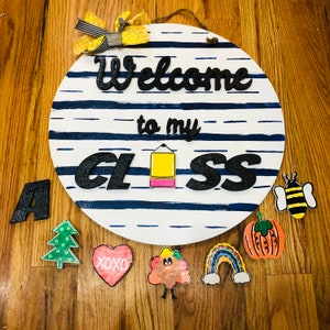 May include: A white round wooden sign with blue and white stripes. The sign says "Welcome to my Class" in black lettering. There are various colourful wooden decorations around the sign, including a bee, a heart, a rainbow, a tree, a pumpkin, and a cloud.