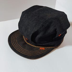 May include: A black denim cap with a brown brim and orange stitching. The cap has a small orange label with the word "ROARK" on it.