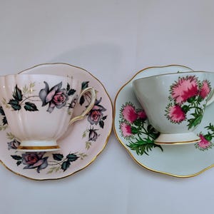 May include: Two vintage teacups and saucers. One is light pink with gray and pink roses, the other is light blue with pink thistle flowers. Both have gold trim around the edges. A classic tea set.