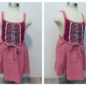 May include: A pink corduroy pinafore dress with a burgundy and gray bodice featuring a floral pattern and lace trim. The dress has adjustable straps and a tie at the waist.