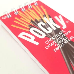 May include: A red spiral notebook with the word "POCKY" in white letters. The notebook features a design of chocolate covered biscuit sticks. The text "CHOCOLATE", "CHOCOLATE CREAM COVERED BISCUIT STICKS" is printed on the notebook.