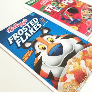 May include: Two cereal boxes: Kellogg's Frosted Flakes with Tony the Tiger and Kellogg's Froot Loops. The Frosted Flakes box is blue with a tiger illustration and the text "THEY'RE GR-R-REAT!". The Froot Loops box is red with the cereal name.