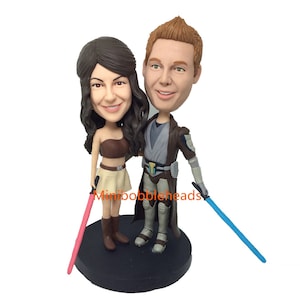 Peut inclure: Un couple de figurines personnalisées habillées en Jedi. La femme tient un sabre laser rose et l'homme un sabre laser bleu. Ils sont debout sur une base noire.