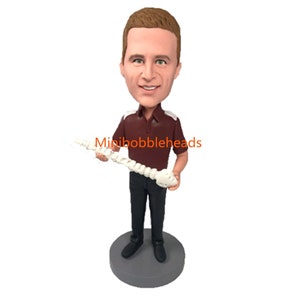 Peut inclure: Une figurine de bobblehead personnalisée d'une personne portant un polo marron avec des bordures blanches et un pantalon noir. La personne tient un modèle blanc de colonne vertébrale humaine. Le bobblehead est sur une base grise. "Minibobbleheads" est imprimé sur la base.