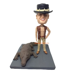 Peut inclure: Une figurine de tête de bobble personnalisée d'une personne portant un gilet en cuir marron, un pantalon kaki et un chapeau noir avec une bande blanche. La personne se tient à côté d'une figurine d'alligator marron.