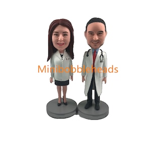 May include: Two custom bobbleheads of doctors wearing white lab coats, one female with brown hair and one male with brown hair. The bobbleheads are standing on grey bases. The text "Minibobbleheads" is visible in the image.