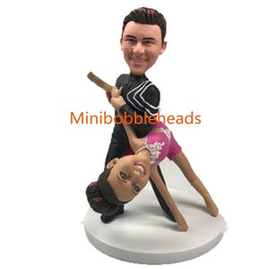 Peut inclure: Une figurine de couple dansant en bobblehead personnalisée. L'homme porte un smoking noir et blanc et la femme une robe rose. La figurine est sur un socle blanc. "Minibobbleheads" est imprimé sur le socle.