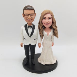 May include: A wedding cake topper featuring a smiling couple. The groom wears a white tuxedo with a black bow tie and glasses. The bride is in a white wedding dress with long sleeves and a plunging neckline. They stand on a black base.