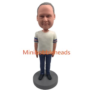 Peut inclure: Une figurine de bobblehead personnalisée représentant une personne aux cheveux gris portant un t-shirt blanc avec des rayures bleues et rouges sur les manches et un jean bleu. La figurine est debout sur une base grise. Le texte "Mini bobbleheads" est visible sur l'image.