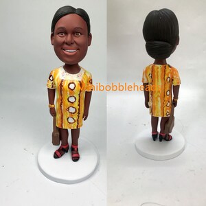 Peut inclure: Une figurine de tête de bobble personnalisée d'une personne portant une robe jaune et orange avec des accents bruns et tenant un sac brun. La figurine est posée sur une base blanche.