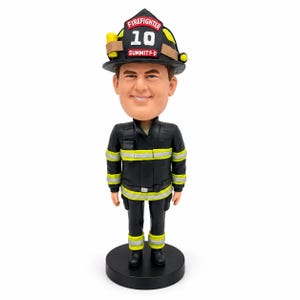 Pode incluir: Uma figura bobblehead de um bombeiro vestindo um uniforme preto com faixas refletoras amarelas e um capacete preto. O capacete apresenta o texto FIREFIGHTER 10 e SUMMIT F-D. A figura tem uma expressão sorridente e está sobre uma base preta.