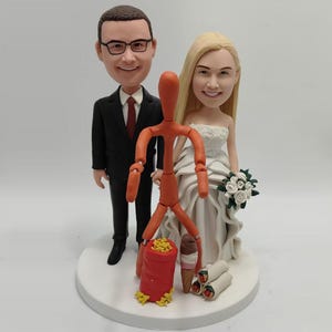 May include: A wedding cake topper featuring a bride and groom figurine with bobbleheads. The groom wears a black suit and the bride a white wedding dress. An orange figurine stands between them, with a red bag of chips, an ice cream cone, and burritos.