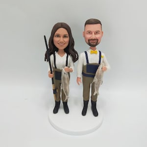 May include: A custom-made figurine couple dressed in fishing gear, holding fish and a fishing rod. The woman is wearing a white shirt and brown pants, while the man is wearing a white shirt, brown pants, and a yellow bow tie.
